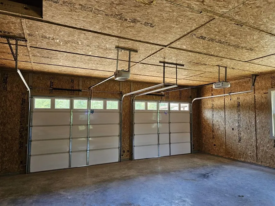 Two white garage doors with window rows and two ceiling-mounted openers in an unfinished garage